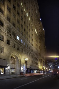 Nighttime low-angle view of a tall stone building on a city street with car light blurs.