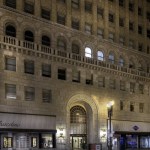 Nighttime low-angle view of a tall stone building on a city street with car light blurs.