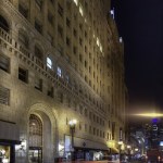 Nighttime low-angle view of a tall stone building on a city street with car light blurs.