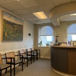 Office waiting area featuring a wooden reception desk, seating, and a large landscape painting.