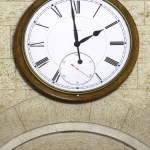 Close-up of a classic analog clock with Roman numerals mounted on a stone wall above an elevator sign.